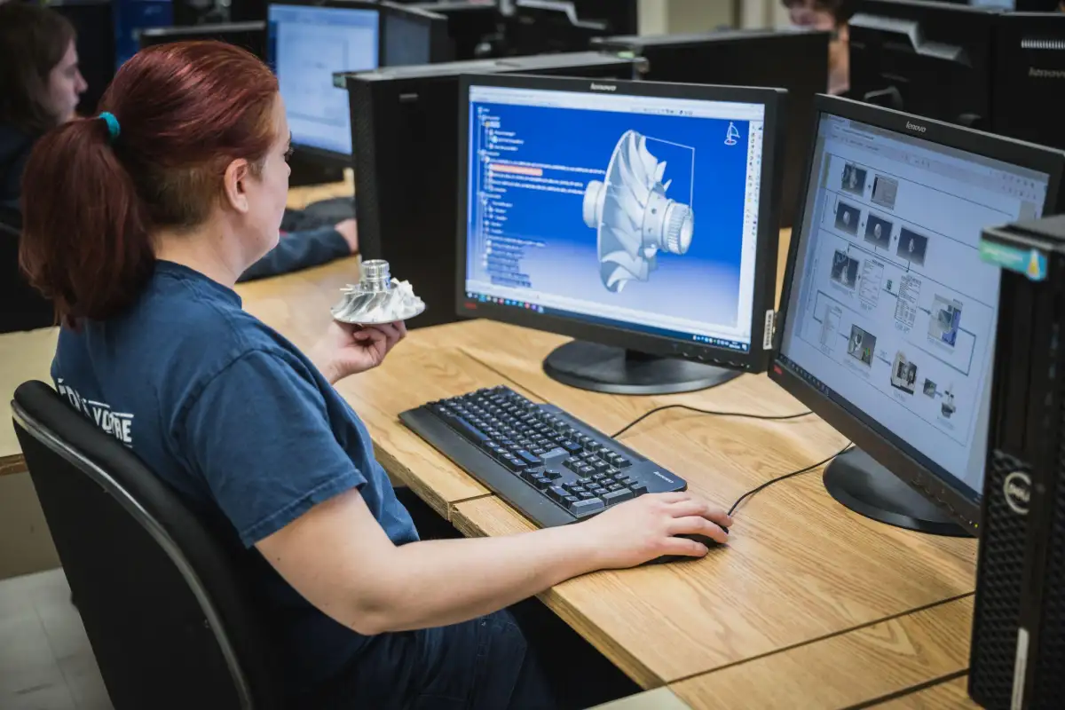 Étudiante dans un laboratoire informatique avec le logiciel CATIA ouvert sur ses écrans et une pièce en métal dans sa main
